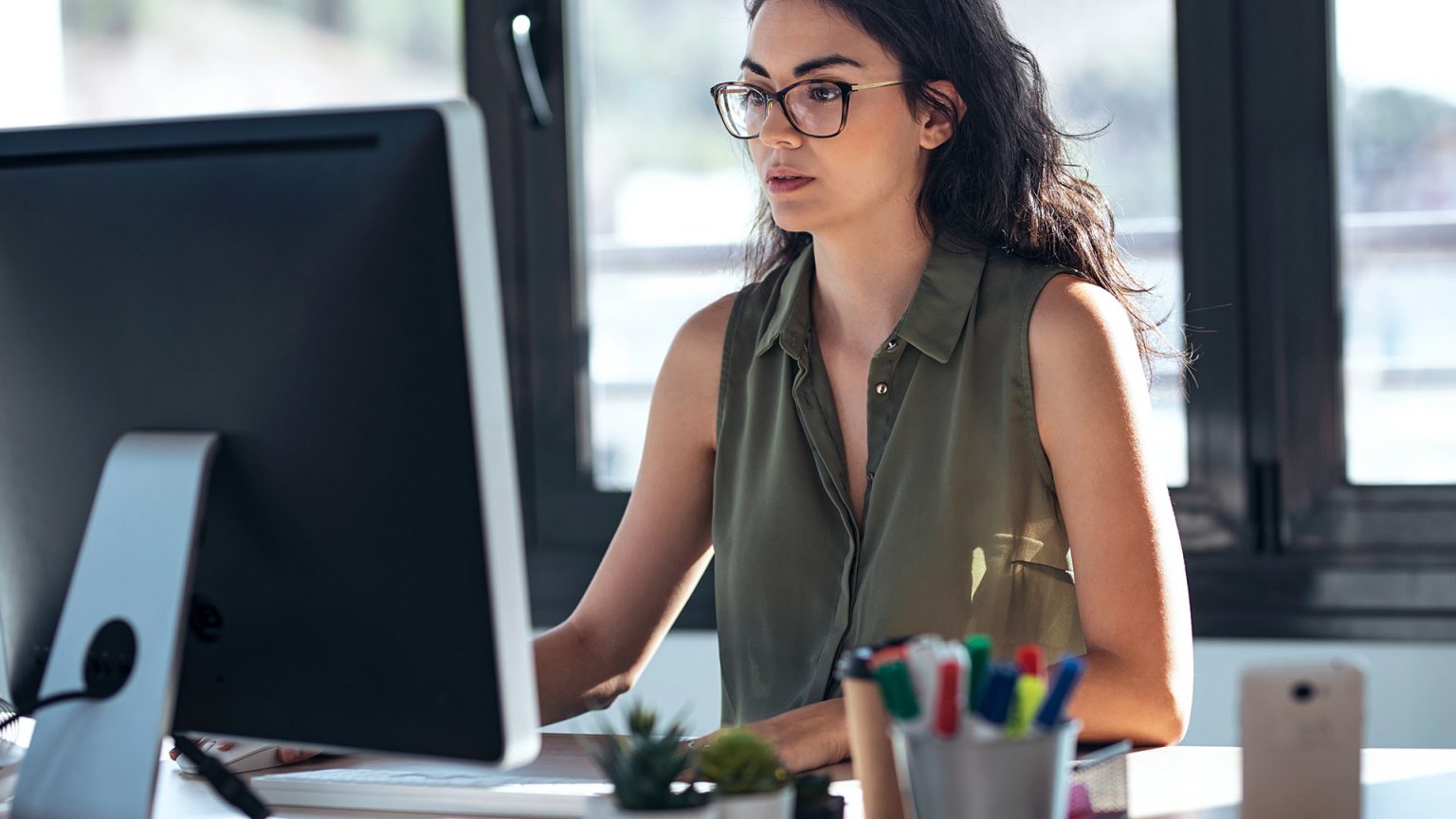  A young woman in glasses sits at her desk looking at her computer screen which displays search results for 'Consequences of deleting YouTube account'.